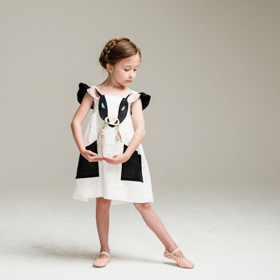 Girl wearing a white cow dress with black appliqué details, flutter sleeves, and a 3D cow face design. Whimsical farm animal outfit.